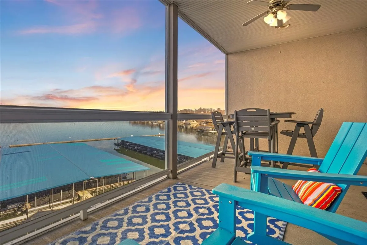 The image showcases a spacious condo balcony overlooking the expansive Lake of the Ozarks main channel. Blue lounge chairs and a dining table are set atop a cozy rug, inviting guests to relax, dine, and take in the breathtaking water views—especially at sunset, when the sky glows over the lake.