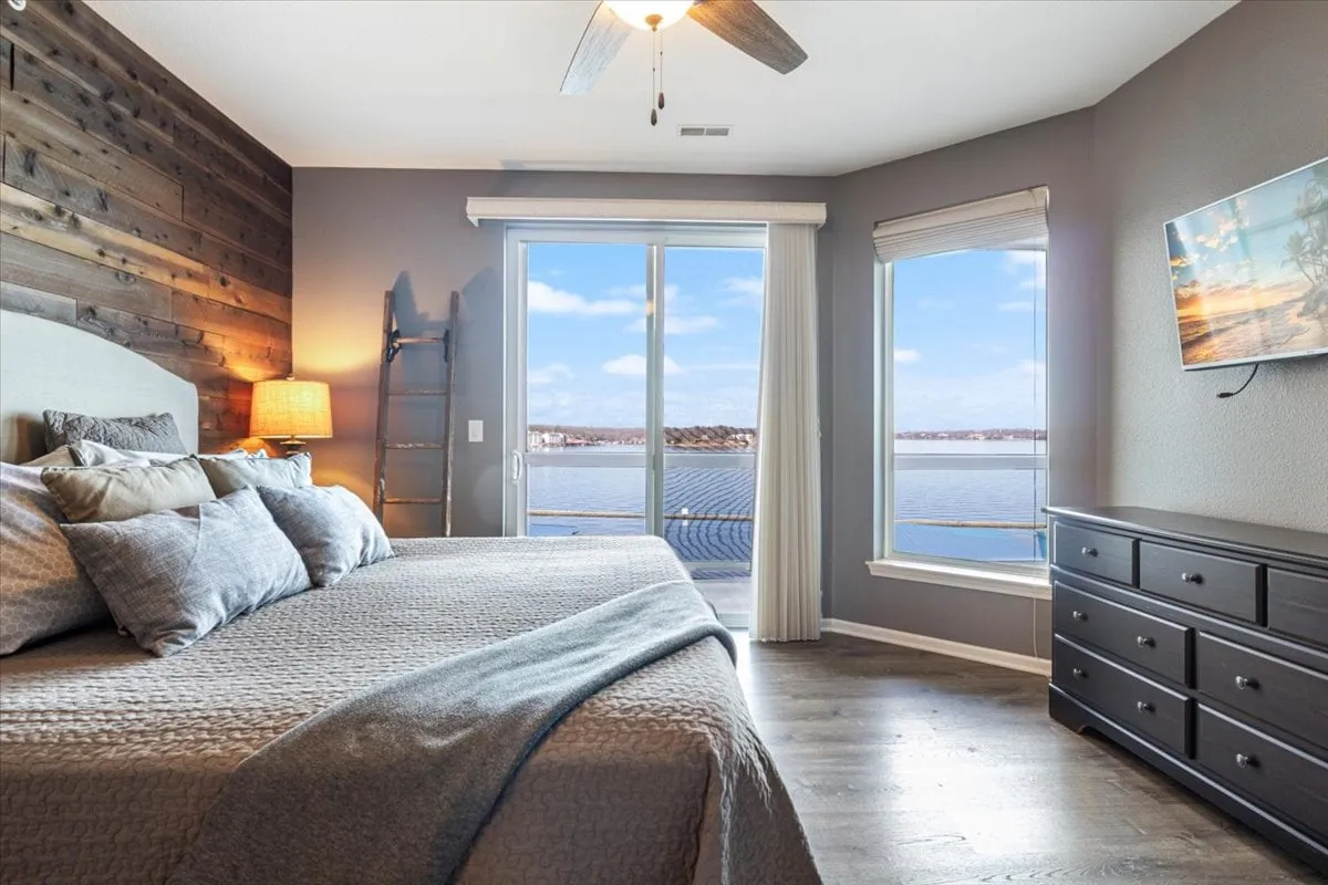 The image showcases a spacious primary bedroom featuring a king-size bed positioned to face large windows that offer a captivating lake view. The design includes a stylish wooden accent wall, a comfortable nightstand with a lamp, and a mounted television, all set in a calm and inviting atmosphere.