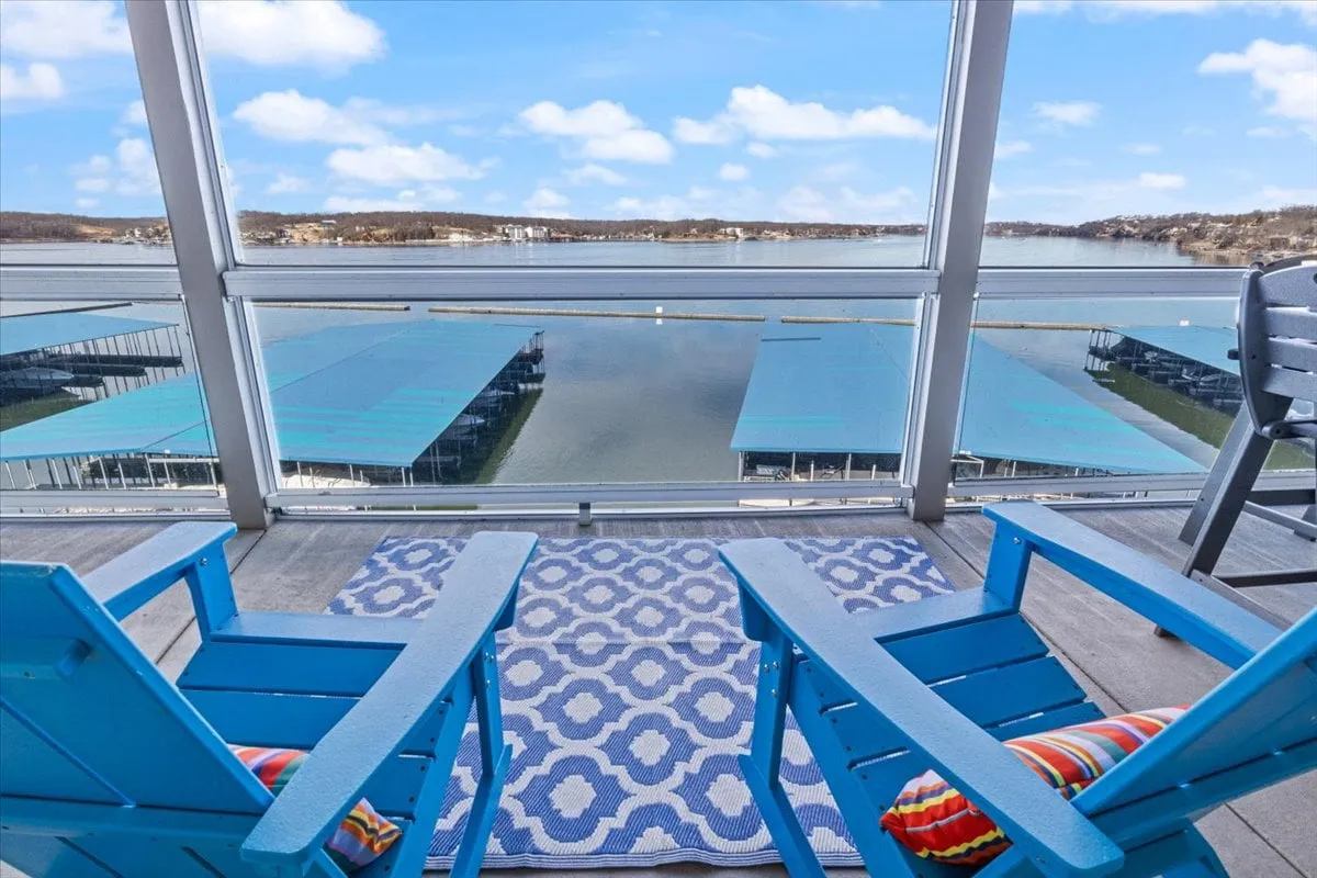A serene outdoor seating area featuring blue chairs and a rug arranged to face a wide, tranquil lake. The inviting arrangement offers a perfect spot for sipping coffee or cocktails while soaking in panoramic water views, with the soft glow of sunset visible on the horizon.