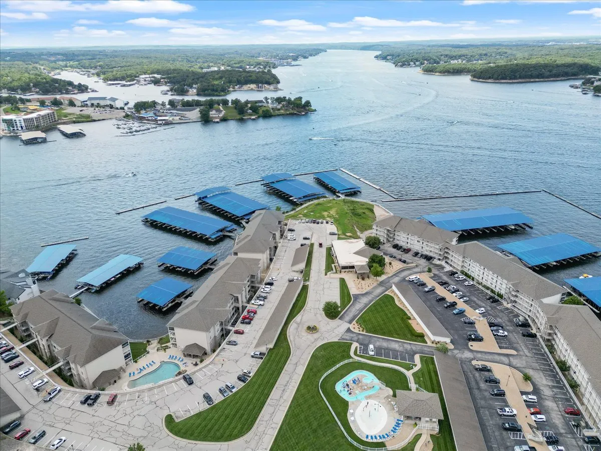 A sweeping exterior view of the property set beside the lake, with the marina, swimming pool, lush green spaces, and residential buildings all visible. The scene invites thoughts of boating, swimming, and lakeside exploration.