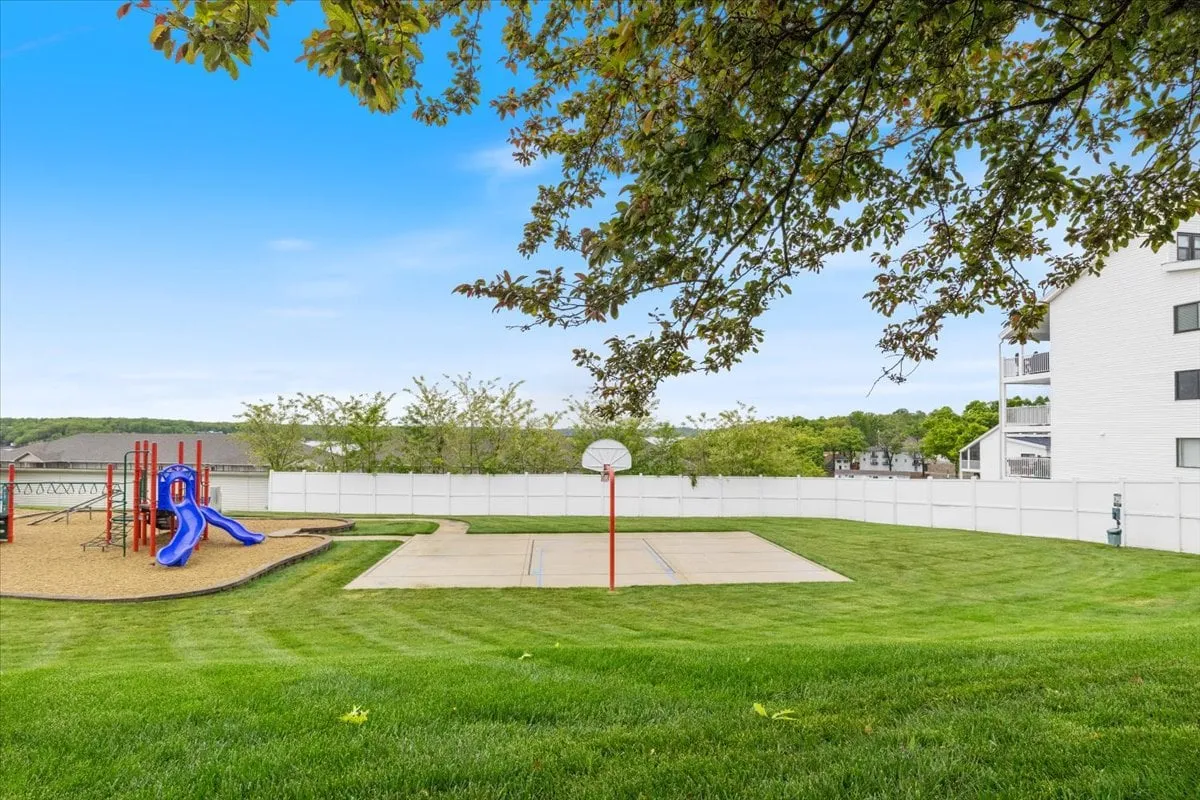 A vibrant scene showing a well-equipped playground structure adjacent to a basketball court. The lush grass and clear blue sky contribute to the energetic, joyful mood, with children playing and families gathered for outdoor activities.