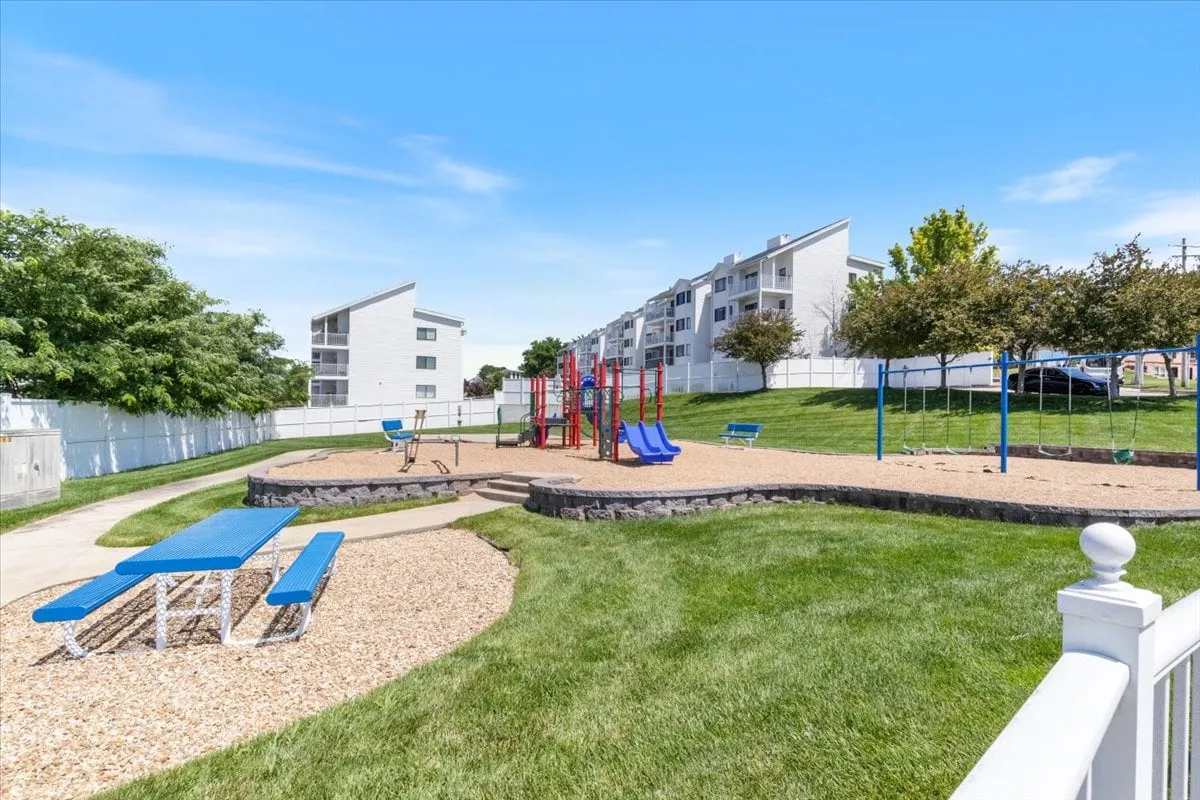 A spacious playground area with modern playground equipment, picnic tables, and green grassy spaces. Children can be seen playing, while picnic tables invite families to gather and enjoy the weather together.