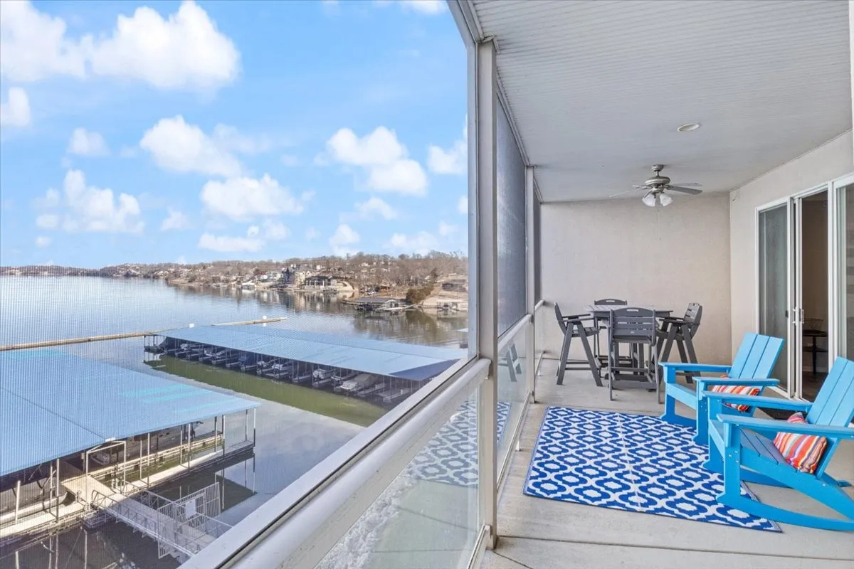 An outdoor balcony features blue chairs and a dining table facing the lake, with a patterned rug adding a touch of comfort. The scene highlights the effortless indoor-outdoor flow and provides a perfect spot for morning coffee, evening barbecues, or simply soaking in the breathtaking lake vistas.